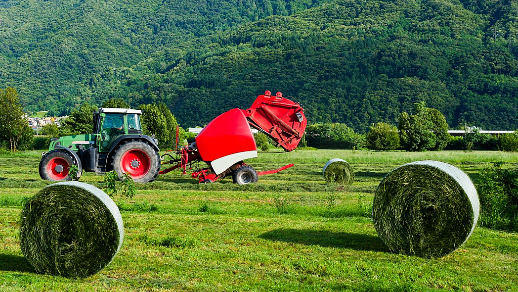 Vorne zwei Rundballen mit Heu, dahinter ein Traktor mit Ballenpresse bei der Arbeit.