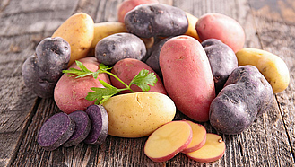 Kartoffeln verschiedener Farben lieben neben und übereinander auf einem Holztisch, einige davon sind in Scheiben geschnitten.