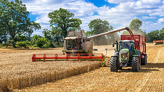 Ein Mähdrescher fährt neben einem Traktor mit Anhänger und befüllt diesen mit Getreide.