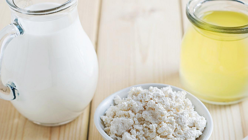 Ein Krug mit Milch, eine Schale mit körnigem Frischkäse und ein Glas mit Molke auf einem Tisch.