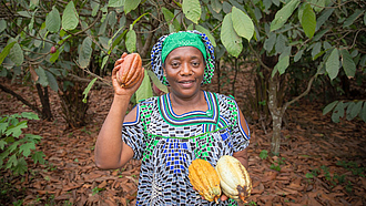 Eine Kamerunerin mit Kakaofrüchten in den Händen steht in einer Kakaoplantage.