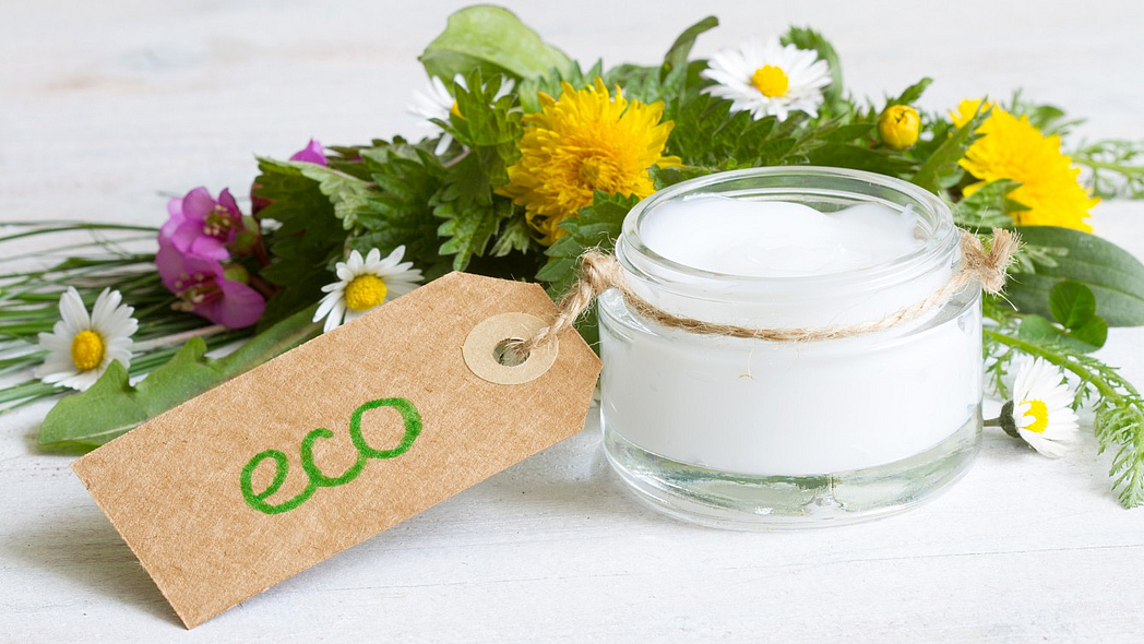 Ein Glas voll Creme, das mit einen Eco-Etikett versehen ist, steht vor einem Arrangement aus Wildblumen.