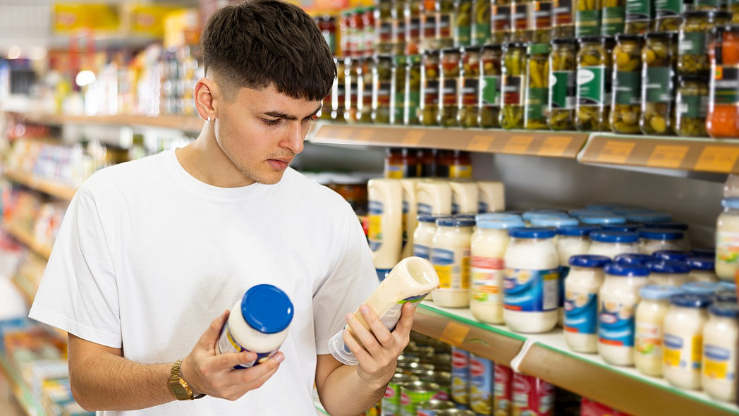 Ein junger Mann vor einem Supermarktregal vergleicht zwei Mayonnaise-Produkte.