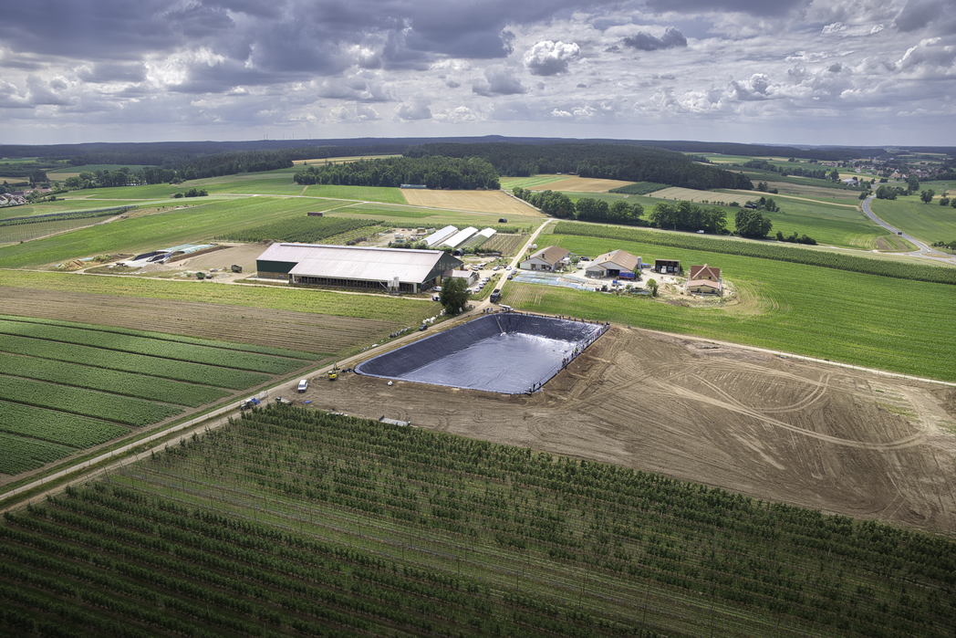 Aus der Vogelperspektive ist ein großes Erdfolienbecken neben einer Obstanlage zu sehen.
