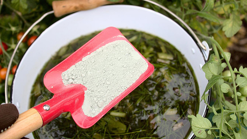 Una pala roja para niños con harina de roca se sostiene sobre un balde con estiércol vegetal.