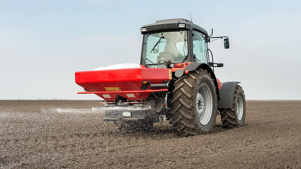 Traktor mit einem roten, angehängtem Mineraldüngerstreuer bei der Arbeit auf dem Acker