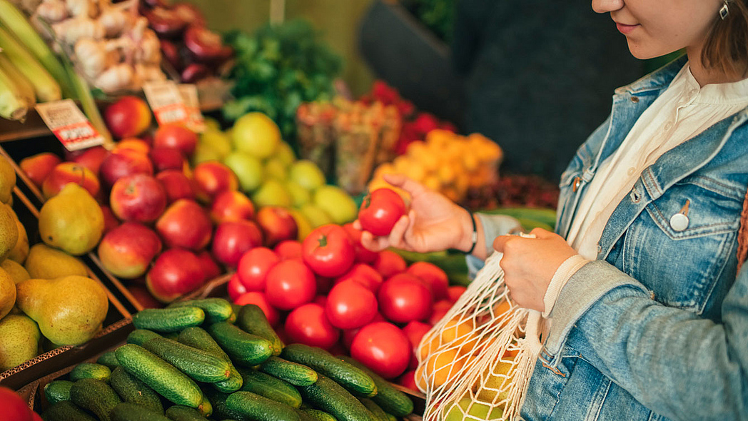 Junge Frau mit Einkaufsnetz vor Obst- und Gemüseregal greift nach einer Tomate. Junge Frau mit Einkaufsnetz vor Obst- und Gemüseregal greift nach einer Tomate.