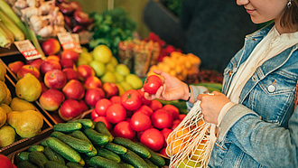 Junge Frau mit Einkaufsnetz vor Obst- und Gemüseregal greift nach einer Tomate.