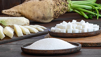 Eine flache Holzschale mit granuliertem Zucker, dahinter eine Holzschale mit Würfelzucker und im Hintergrund eine Zuckerrübe