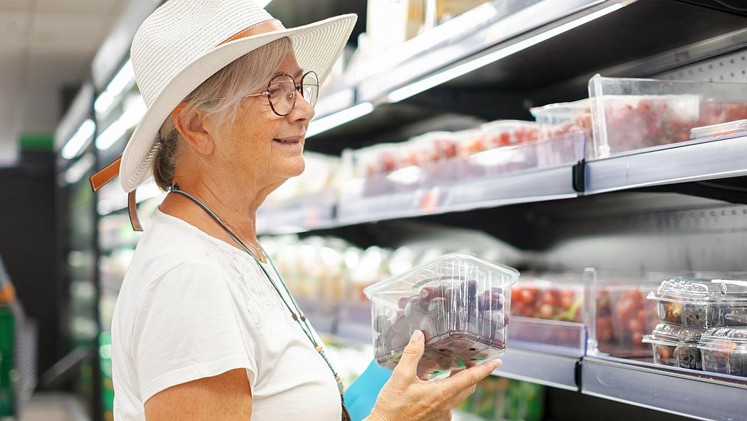 Eine ältere Frau mit weißem Hut steht in einem Supermarkt vor einem Regal mit Lebensmitteln. In der Hand hält sie eine Schale mit Trauben.