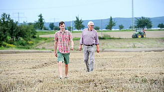 Ein jüngerer und ein älterer Landwirt gehen über ein Feld.
