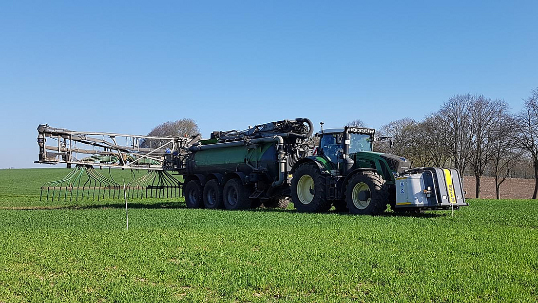 Traktor bei der Ausbringung von Gülle auf einer Wiese. Vorne am Traktor ist ein Säurefass montiert.