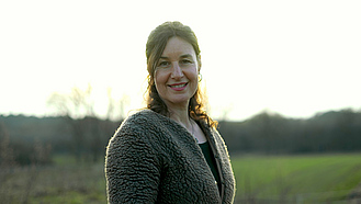 Eine Frau mit langen, dunklen Haaren steht auf einer Wiese vor einem hellen Himmel.