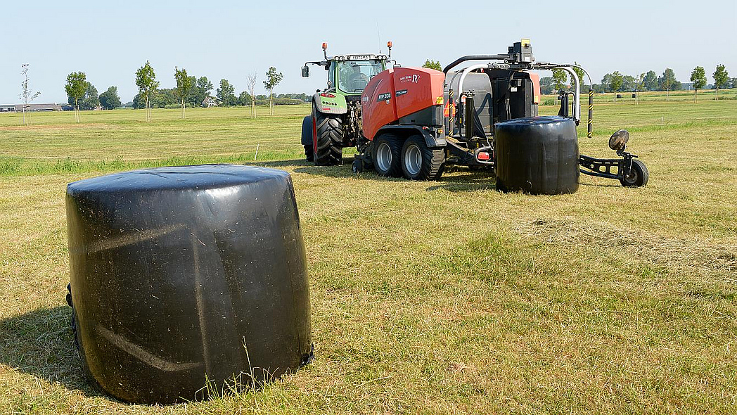 In Folie gewickelter Rundballen; im Hintergrund eine Rundballenpresse